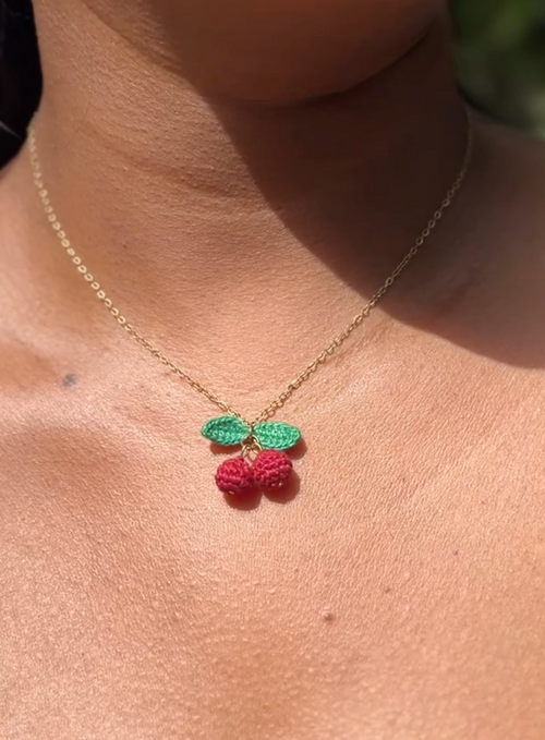 A close-up of a person wearing a delicate gold chain necklace with a crocheted cherry pendant featuring green leaves and red cherries.